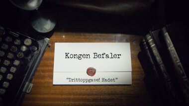 Image of the title card at the start of this episode, showing a task brief with the show title, 'Kongen Befaler', and the episode title, 'Drittoppgave! Hadet' ['Shit task! Goodbye'], on a wooden desk. At the edges of the image, part of a typewriter keyboard and a stack of leather-bound books can be seen.