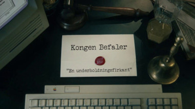 Image of the title card at the start of this episode, showing a task brief with the show title, 'Kongen Befaler', and the episode title, 'En underholdningsfirkant' ['An entertainment square'], on a wooden desk. Also on the desk is an old-fashioned Apple computer and keyboard, a gavel, an hourglass, and a bell.