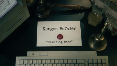 Image of the title card at the start of this episode, showing a task brief with the show title, 'Kongen Befaler', and the episode title, 'Fakk slag, mann' ['Fuck strikes, man'], on a wooden desk. Also on the desk is an old-fashioned Apple computer and keyboard, a gavel, an hourglass, and a bell.