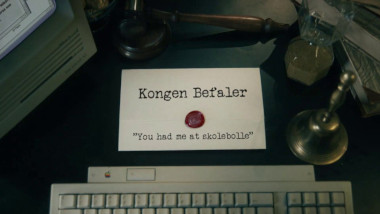 Image of the title card at the start of this episode, showing a task brief with the show title, 'Kongen Befaler', and the episode title, 'You had me at skolebolle' ['You had me at school bun'], on a wooden desk. Also on the desk is an old-fashioned Apple computer and keyboard, a gavel, an hourglass, and a bell.