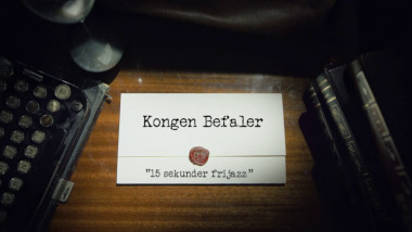 Image of the title card at the start of this episode, showing a task brief with the show title, 'Kongen Befaler', and the episode title, '15 sekunder frijazz' ['15 seconds of free-jazz'], on a wooden desk. At the edges of the image, part of a typewriter keyboard and a stack of leather-bound books can be seen.