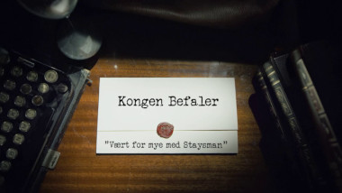 Image of the title card at the start of this episode, showing a task brief with the show title, 'Kongen Befaler', and the episode title, 'Vært for mye med Staysman' ['Been spending too much time with Staysman'], on a wooden desk. At the edges of the image, part of a typewriter keyboard and a stack of leather-bound books can be seen.