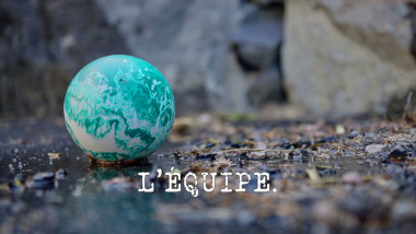 Image of a small blue-green bowling ball on some wet, rocky ground (a reference to Phil Roy's attempt at the 'Make the highest splash' task), with the episode title, 'L'ÉQUIPE' ['THE TEAM'], superimposed on it.