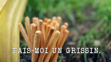 Close-up image of some breadsticks next to the edge of a yellow plastic slide (a reference to the ‘Push a watermelon up a slide using breadsticks’ task), with the episode title 'Fais-moi un Grissin' ['Make Me a Breadstick'] superimposed on it.