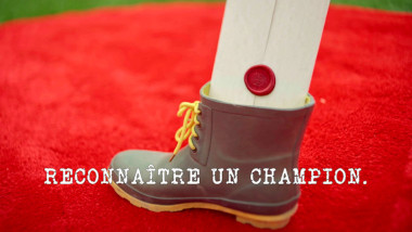 Image of a task brief stuck into a work boot on a red rug (a reference to the ‘Catapult a boot into a bathtub’ task), with the episode title 'Reconnaître un champion' ['Recognise a champion'] superimposed on it.