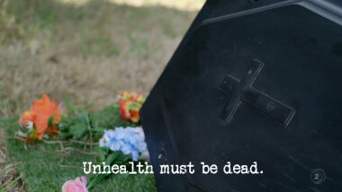 Image of an open coffin lid, with flowers behind it (a reference to Angella's attempt at the 'Be as unhealthy as possible' task), with the episode title, 'Unhealth must be dead', superimposed on it.