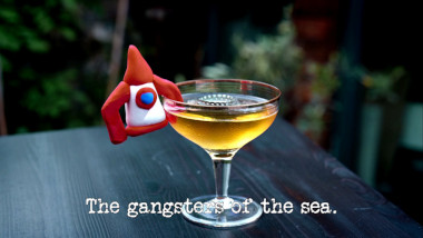 Photo of a red and white modelling clay rocket, hung over the side of a cocktail glass full of an amber-coloured beverage, and with a bottle cap floating on the surface (referencing the 'Put a rocket in your pocket' and 'Win the pub quiz by cheating' tasks), with the episode title, 'The gangsters of the sea', superimposed on it.