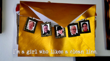 Photo of a golden envelope, with miniature framed images of all of the contestants strung across it (referencing the 'Push the envelope the furthest' and 'Pick the Taskmaster’s locket from its pocket' tasks), with the episode title, 'I'm a girl who likes a clean line', superimposed on it.