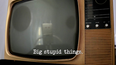 Photo of the screen of an old-fashioned CRT television, with the reflection of a robot on it (referencing the 'Direct the robot to its charging station' and 'Present the most heart-warming final local news story' tasks), with the episode title, 'Big stupid things', superimposed on it.