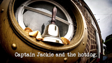 Photo of a handbell and some fortune cookies resting in the porthole frame of one of the round windows at the front of the Taskmaster house (referencing the 'Ring the bell' and 'Eat and obey five fortune cookies' tasks), with the episode title, 'Captain Jackie and the hotdog', superimposed on it.