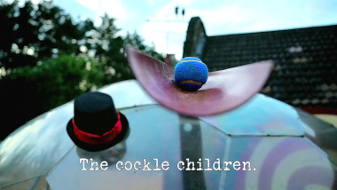 Photo of a blue tennis ball in a length of red guttering, and a top hat with a ribbon around it, both balanced on the geodesic dome (referencing the 'Walk exactly one furlong in exactly 10 minutes' and 'Place all the balls in the gutter, on the cow' tasks), with the episode title, 'The cockle children', superimposed on it.