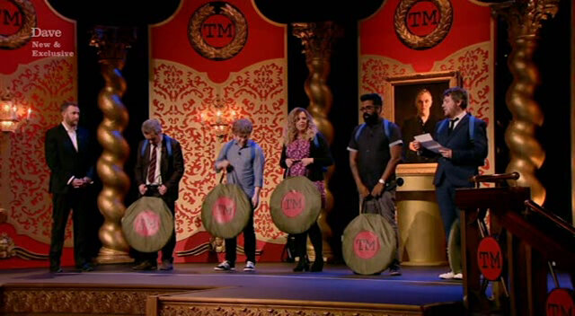The cast of Taskmaster UK series 1 standing on stage wearing rucksacks and holding collapsed pop-up tents as Tim Key reads out the task brief. The cast of Taskmaster UK series 1 standing on stage wearing rucksacks and holding collapsed pop-up tents as Tim Key reads out the task brief.