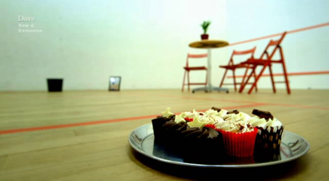 Image of a squash court, with a tray of cupcakes in the foreground, and a waste paper bin, framed picture, and table (with potted plant) and chairs in the background. Image of a squash court, with a tray of cupcakes in the foreground, and a waste paper bin, framed picture, and table (with potted plant) and chairs in the background.