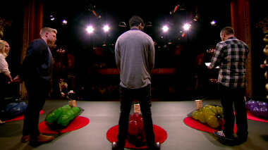 Dave Gorman reads out a task as the cast of Series 3 stands looking at black mesh bags filled with inflated balloons.