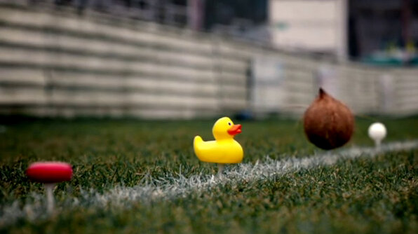 Four items are set atop golf tees along a white line in the grass: A small round cheese, a rubber duck, a coconut, and a ping-pong ball. Four items are set atop golf tees along a white line in the grass: A small round cheese, a rubber duck, a coconut, and a ping-pong ball.