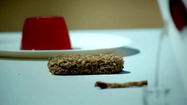 A white table with the following items: Jelly on a plate, a Twiglet, a Weetabix and a partially visible sealed task brief.