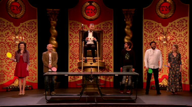 Alex Horne sitting on a referees chair behind a ping pong table in the Studio holds out a sealed task brief. The cast of UK series 5 is lined up next to him holding ping-pong paddles in their paddles.