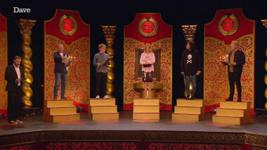 The cast of UK Champion of Champions 1 on the Taskmaster studio stage. Each contestant stands on a set of stairs three steps high holding a whiteboard and marker. Alex stands off to the side with his clipboard as Bob Mortimer reads out the task.