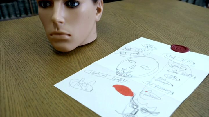 A mannequin's head rests on the living room table next to an open task brief lying face down. Some masks and a list of ingredients are scribbled on the back of the task. A mannequin's head rests on the living room table next to an open task brief lying face down. Some masks and a list of ingredients are scribbled on the back of the task.