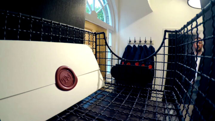 A sealed task brief resting in a wire basket in a Taskmaster UK house pigeon-hole. Alex Horne stands in the background. Five boiler suits are visible on coat hooks next to the front door. A sealed task brief resting in a wire basket in a Taskmaster UK house pigeon-hole. Alex Horne stands in the background. Five boiler suits are visible on coat hooks next to the front door.