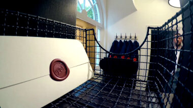 A sealed task brief resting in a wire basket in a Taskmaster UK house pigeon-hole. Alex Horne stands in the background. Five boiler suits are visible on coat hooks next to the front door.