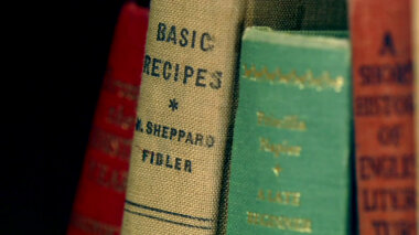 Four vintage cloth-bound books on a shelf. The legible title is Basic Recipes.