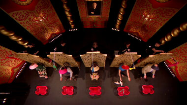 Aerial view of the Taskmaster UK studio stage. The Taskmaster UK series 6 cast sit on chairs, each in front of a barrier. They are wearing visors and have red laundry baskets in front of them. Behind each barrier, a crew member dressed in black is standing. They each hold a grabbing and reaching tool and are dangling a sock over the head of the contestant in front of them.