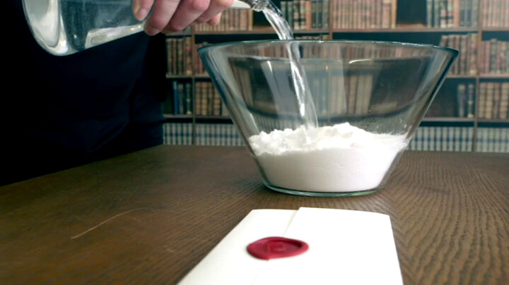 A glass bowl filled with white powder is on the living room table next to a sealed task brief. Alex Horne is pouring a glass carafe of water into the white powder. A glass bowl filled with white powder is on the living room table next to a sealed task brief. Alex Horne is pouring a glass carafe of water into the white powder.