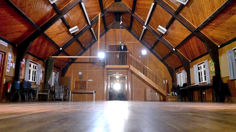 Interior of an A-frame building. Alex Horne stands at the top of a set of stairs holding a contraption which will release a sealed task brief attached to a parachute. Interior of an A-frame building. Alex Horne stands at the top of a set of stairs holding a contraption which will release a sealed task brief attached to a parachute.