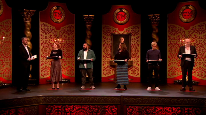 The cast of Taskmaster UK series 6 and Alex Horne stand on the Taskmaster studio stage. Each contestant is standing on a red dot and holding a white board and marker. Alex holds a sealed task brief. The cast of Taskmaster UK series 6 and Alex Horne stand on the Taskmaster studio stage. Each contestant is standing on a red dot and holding a white board and marker. Alex holds a sealed task brief.