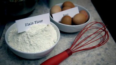 A bowl of flour labelled 'Plain Flour', a bowl of eggs labelled 'Eggs' and a whisk resting on a countertop.
