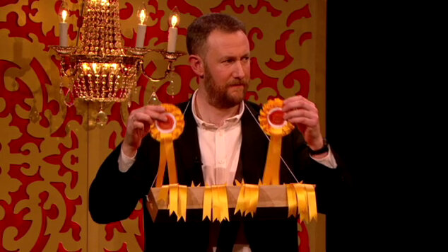 Alex Horne wears a cigarette-girl style tray full of yellow award rosettes. He holds one rosette in each hand. Alex Horne wears a cigarette-girl style tray full of yellow award rosettes. He holds one rosette in each hand.