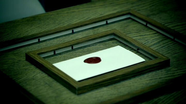 A wooden table. On the table is a wooden picture frame. Inside that is a smaller wooden picture frame. Inside that is a sealed task. A wooden table. On the table is a wooden picture frame. Inside that is a smaller wooden picture frame. Inside that is a sealed task.