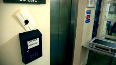 A black mailbox with a sign that says 'feedback welcome'. A sealed task is stuck in behind the top of the box. Next to the box is a set of elevator doors. There is also a small metal table with a closed silver and black briefcase on top.