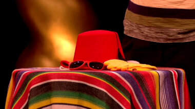 A brightly coloured striped blanket draped over a table. On top is a red fez, red plastic sunglasses, and yellow rubber gloves.
