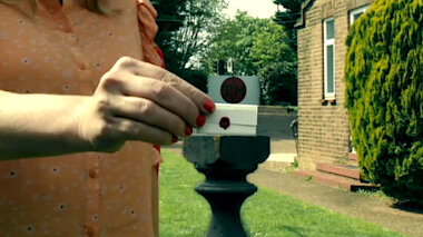 A close-up of Kerry Godliman's right arm and hand. She is picking up a very tiny sealed task from a black pedestal in the Taskmaster house garden.