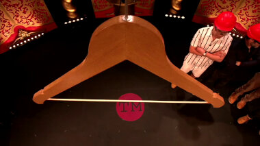 A close-up of a wooden hanger above the Taskmaster studio stage. Phil Wang and Rhod Gilbert can be seen below and to the right of the hanger. They are wearing red plastic hard hats.