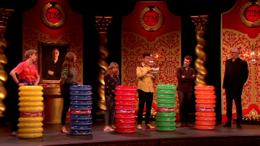 The contestants and Greg Davies on the Taskmaster studio stage. In front of each contestant is a stack of inflatable pool rings, aka doughnuts. Phil Wang is reading the task.