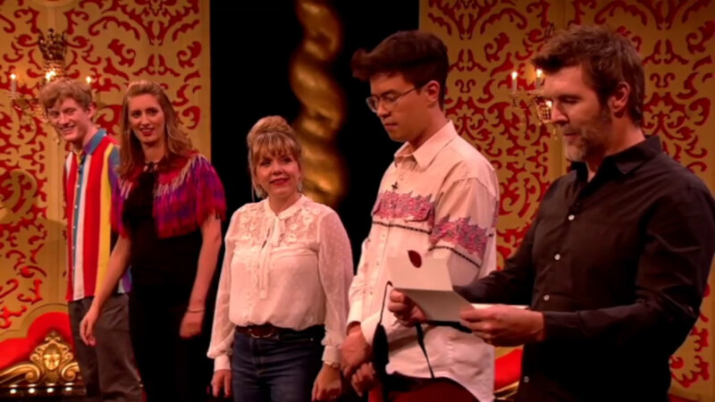 All five contestants on the Taskmaster studio stage. Four of them are watching Rhod Gilbert read the final task of the episode. All five contestants on the Taskmaster studio stage. Four of them are watching Rhod Gilbert read the final task of the episode.