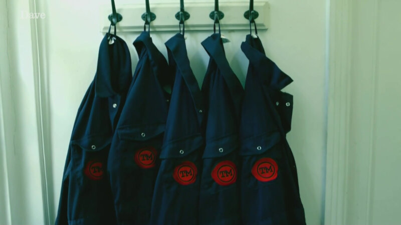 Five pairs of blue coveralls hanging on hooks. Each pair bears the Taskmaster logo. Five pairs of blue coveralls hanging on hooks. Each pair bears the Taskmaster logo.