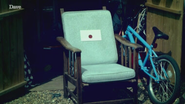 A wooden chair with green cushions next to a blue bicycle. Attached to the chair cushions is a sealed task.
