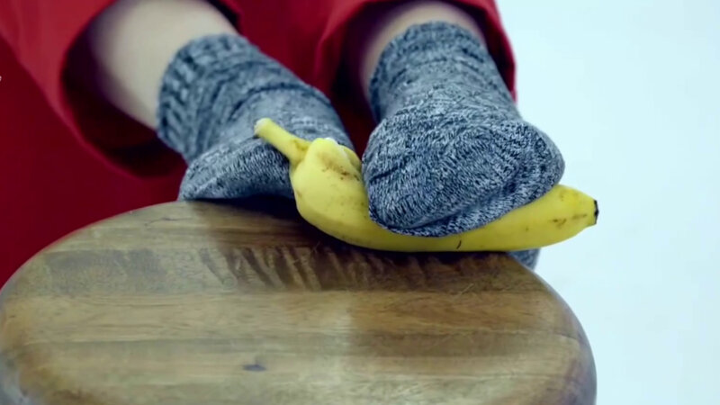 A close-up of Lou Sanders holding a banana between her feet which are resting on a wooden stool. She is wearing red trousers and grey mottled socks. A close-up of Lou Sanders holding a banana between her feet which are resting on a wooden stool. She is wearing red trousers and grey mottled socks.