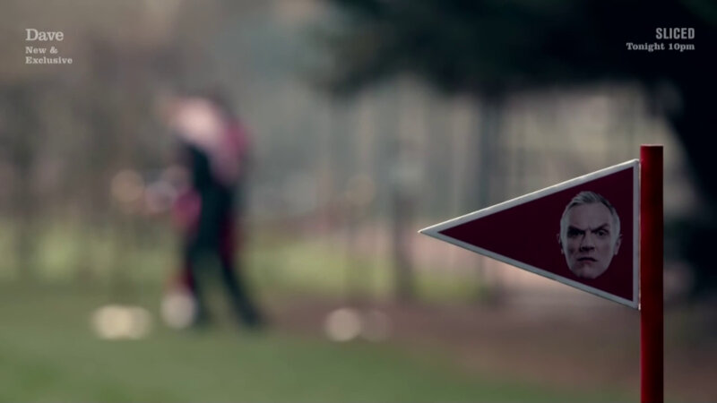 A close-up of a red and white triangular marker flag. On the flag is a photo of Greg Davies. An out of focus person is in the background. A close-up of a red and white triangular marker flag. On the flag is a photo of Greg Davies. An out of focus person is in the background.