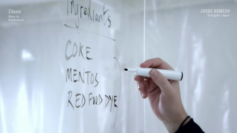 A close-up of Iain Stirling's hand. He is writing a list on plexiglass with a black marker. The list reads 'Ingredients, Coke, Mentos, red food dye.' A close-up of Iain Stirling's hand. He is writing a list on plexiglass with a black marker. The list reads 'Ingredients, Coke, Mentos, red food dye.'