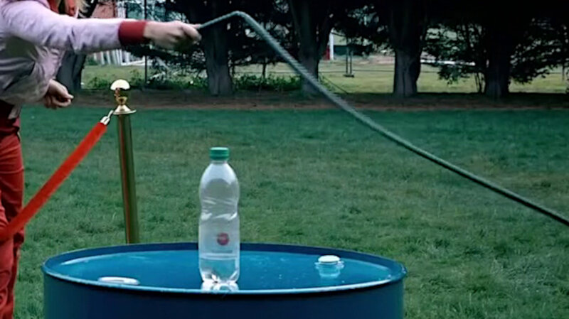 A close-up of a blue metal barrel with a plastic water bottle on top. Part of Lou Sanders can be seen behind a red stanchion. She is holding a whip. A close-up of a blue metal barrel with a plastic water bottle on top. Part of Lou Sanders can be seen behind a red stanchion. She is holding a whip.