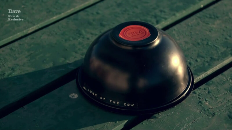 A close-up of a black bowl on a green picnic table. There is a Taskmaster logo on the bowl and the words 'look at the cow'. A close-up of a black bowl on a green picnic table. There is a Taskmaster logo on the bowl and the words 'look at the cow'.