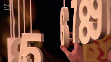 A sequence of white numbers hanging on string. The numbers 5, 6, 7, 8 and 9 are clearly seen. A hand is touching the number 6.