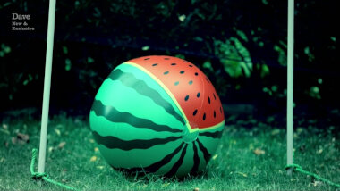 A beach ball that looks like a watermelon sitting on a lawn between two wooden sticks.