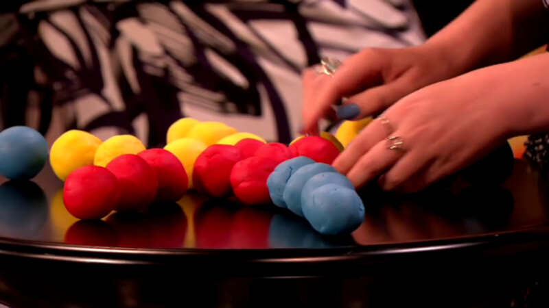 A small round black table covered in yellow, red and blue playdough balls. Katy Wix's hands are starting to pick up one of the red balls. A small round black table covered in yellow, red and blue playdough balls. Katy Wix's hands are starting to pick up one of the red balls.