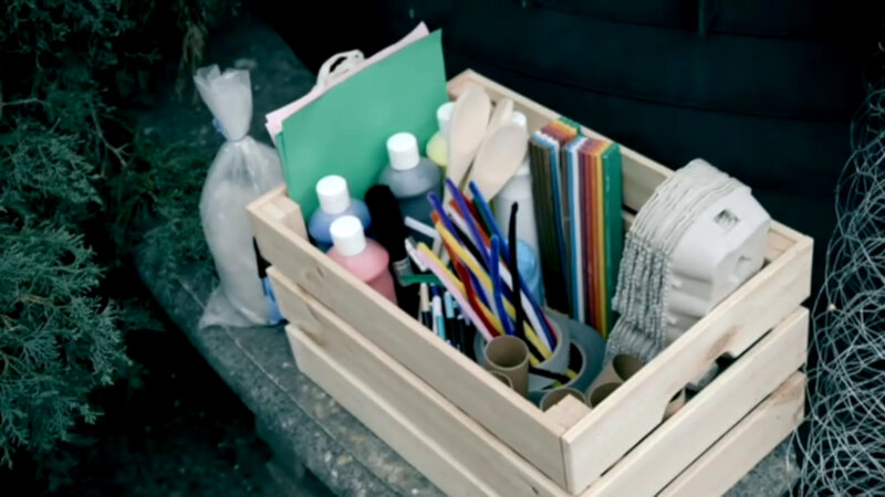 A wooden crate filled with craft supplies including pipe cleaners, bottles of paint, wooden spoons, construction paper, egg cartons, colouring pencils, and cardboard tubes. A wooden crate filled with craft supplies including pipe cleaners, bottles of paint, wooden spoons, construction paper, egg cartons, colouring pencils, and cardboard tubes.